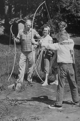 a child shooting a bow and arrow with three adults watching b&w