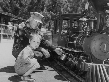 adult dressed as a conductor and child crouching beside a small train b&w