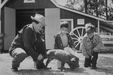 one adult and two children with a small pig in front of the barn b&w
